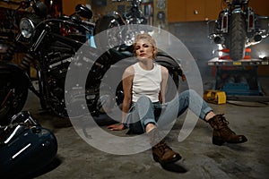 Pretty trendy girl mechanic sitting on garage floor nearby motorcycle