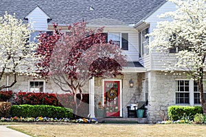 Pretty two story house with rock bottom and white frame with shutters on top in springtime with pansies and flowering trees and