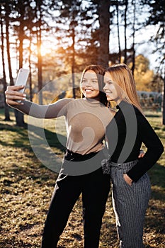 Pretty two young twin sisters in casual outfit with bright smile take selfie with smartphone at the park