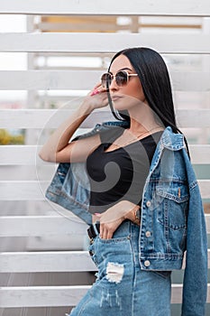 Pretty urban young woman fashion model with luxurious black hair in trendy blue denim clothes in stylish dark sunglasses