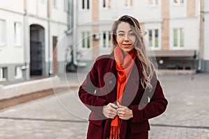 Pretty urban young woman in spring fashionable burgundy coat with stylish orange scarf posing on street. Trendy attractive girl