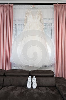 Pretty wedding white dress hanging at hme with white wedding shoes on black sofa, vertical shot