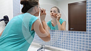 Pretty woman applying black mascara for eyelashes makeup in front of bathroom mirror