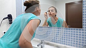 Pretty woman applying black mascara for eyelashes makeup in front of bathroom mirror