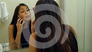 Pretty woman applying mascara eyelash in front of the mirror, while talking via smartphone in bathroom