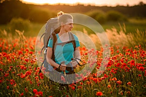 Pretty woman with backpack and walking sticks stands on field of poppies and looks away