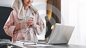 Pretty woman in bathrobe using laptop at table with partner in background at home in the kitchen