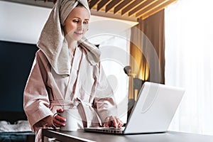 Pretty woman in bathrobe using laptop at table with partner in background at home in the kitchen