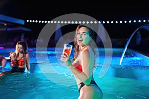 Pretty woman in bikini drinking cocktail and hanging out with friends at night pool party. People having fun dancing