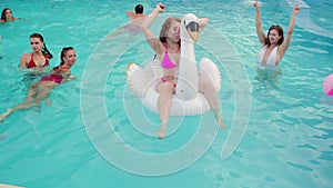 Pretty woman in bikini swimsuit hanging out on inflatable white swan mattress on night pool party. Friends partying with