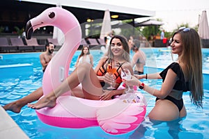 Pretty woman in bikini swimwear hanging out on inflatable pink flamingo mattress on night pool party. Friends partying