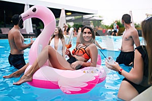 Pretty woman in bikini swimwear hanging out on inflatable pink flamingo mattress on night pool party. Friends partying