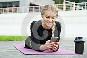 Pretty woman with blond hair lying on a fitness rug with smartphone in hands. Chatting after training exercise