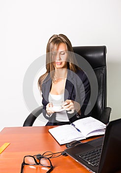 Pretty woman in a business suit sitting at a desk with computer. woman drinks coffee from a white cup