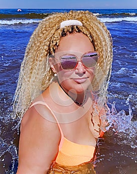 Pretty woman with curly hair in a bright swimsuit and sunglasses splashing in the waves of the Baltic Sea in Jurmala, Latvia.