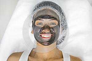 pretty woman with a face mask lying on a table and smiling to the camera, closeup spa center beauty treatment concept