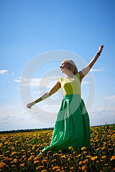 Pretty woman on a field with green grass and yellow dandelion flowers in a sunny day. Girl on nature with yellow flowers and blue