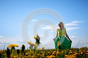 Pretty woman on a field with green grass and yellow dandelion flowers in a sunny day. Girl on nature with yellow flowers and blue