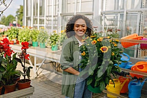 Pretty woman gardener holding flower pot while standing on greenhouse yard background