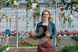 pretty woman gardener looking at camera holding notebook make notes, in greenhouse.