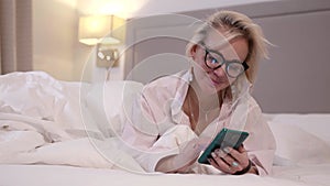 Pretty woman with glasses is lying in bed at evening and chatting in dating app in smartphone