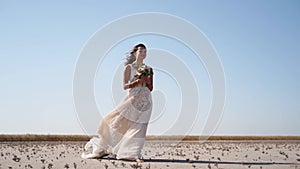 Pretty woman in gorgeous wedding dress walking at desert landscape, windy weather. Elegance bride with bouquet of fresh