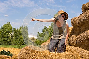 Pretty woman with hat standing in straw bales and pointing her finger toward the blue sky.