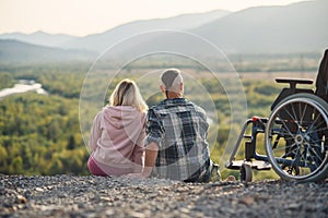 Pretty woman and her incapacitated husband resting together near his wheelchair on the hill.