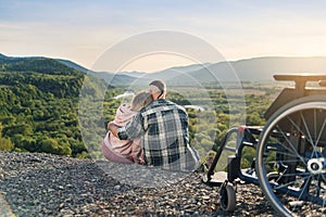 Pretty woman and her incapacitated husband resting together near his wheelchair on the hill.