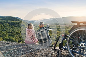 Pretty woman and her incapacitated husband resting together near his wheelchair on the hill.