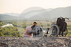 Pretty woman and her incapacitated husband resting together near his wheelchair on the hill.