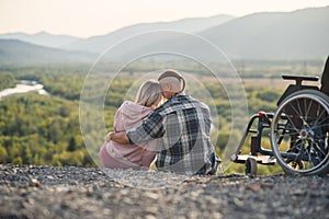 Pretty woman and her incapacitated husband resting together near his wheelchair on the hill.