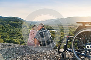 Pretty woman and her incapacitated husband sit near his wheelchair on the hill and kissing gently.