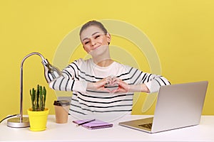 Pretty woman with kind smile sitting at workplace, showing romantic heart shape with hand.