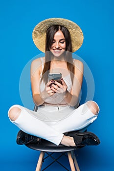 Pretty woman with long hair in hat chilling in chair on blue background and typing on phone.