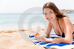 Pretty woman is lying on the towel at the beach and opening a tube of sun cream to protect her skin from sunburn