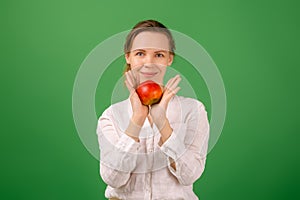 A pretty woman of middle age in a white shirt holds an apple in her hand on a green background. Healthy eating, vegetarianism,