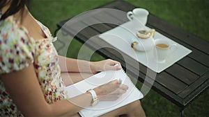Pretty woman in a nightie has breakfast outside and writes a letter on the paper on her lap.