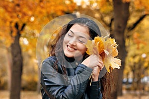 Pretty woman is posing with bunch of maple`s leaves in autumn park. She`s closing eyes and dreaming. Beautiful landscape at fall