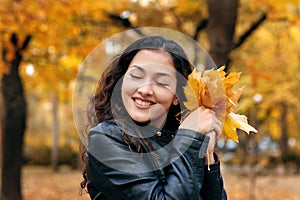 Pretty woman is posing with bunch of maple`s leaves in autumn park. She`s closing eyes and dreaming. Beautiful landscape at fall