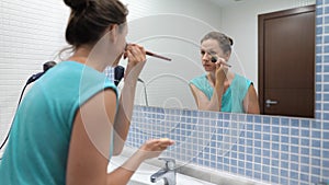 Pretty woman powdering face with makeup brush in front of bathroom mirror