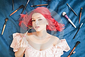 Pretty woman with red hair lying with make up brushes and dreaming, top view girl make up master on blue background