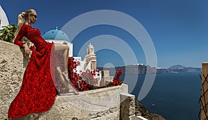 Pretty woman in red long evening dress is posing in Oia, Santorini Island. Female model in amazing long dress. Couture