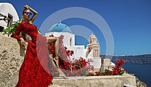 Pretty woman in red long evening dress is posing in Oia, Santorini Island. Female model in amazing long dress. Couture