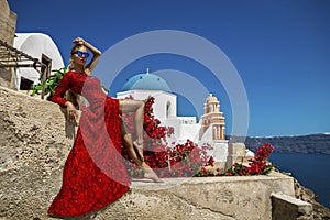 Pretty woman in red long evening dress is posing in Oia, Santorini Island. Female model in amazing long dress. Couture