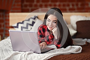 Pretty woman relaxing on the bed in her room and chatting on her laptop. Indoors. Online friendship and social network