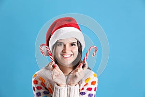 Pretty woman in Santa hat and festive sweater holding candy canes on light blue background