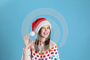 Pretty woman in Santa hat and festive sweater showing OK gesture on light blue background