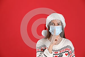 Pretty woman in Santa hat and medical mask showing silence gesture on red background, space for text