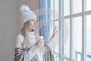 Pretty woman in season casual clothes white sweater and knitted hat holding warm drink indoor